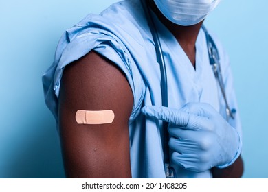 Close Up Of Bandaged Vaccine Shot On Arm Of Medical Assistant Over Blue Background. Healthcare Nurse With Gloves Pointing At Vaccination Plaster For Covid 19 Protection And Prevention