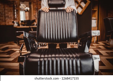 Close Up Barber Leaned Back Against Black Leather Chair In Barbershop. Brown Brick Wall Background. Loft Style