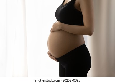 Close Up Of Bare Baby Bump With Good Smooth Skin. Young Pregnant Woman Holding, Caressing Big Belly, Standing By Window, Feeling Kicks Of Unborn Child. Cropped Shot, Close Up. Pregnancy Concept