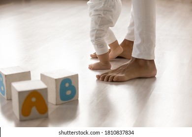 Close Up Barefoot African American Woman Teaching Little Biracial Baby Kid Walking On Warm Wooden Floor Indoors. Happy Mixed Race Family Involved In Daycare Activity, Making First Steps In Living Room