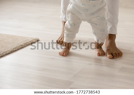 Close up barefoot African American woman with toddler child standing on warm wooden floor with underfloor heating, caring loving young mum teaching adorable little daughter to walk at home