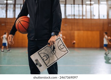 Close up of basketball trainer's hands holding ball and clipboard on training at indoor court. An unrecognizable basketball coach standing at court on training and holding ball and clipboard in hands. - Powered by Shutterstock