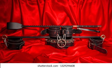 Close Up Bdsm Outfit. Bondage, Kinky Adult Sex Games, Kink Lifestyle Concept With A Pair Of Leather Handcuffs, Flogger, Whip On Red Silk With Copy Space.