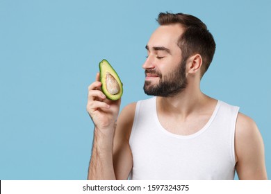 Close Up Bearded Young Man 20s Years Old In White Shirt Hold Half Of Avocado Isolated On Blue Pastel Background, Studio Portrait. Skin Care Healthcare Cosmetic Procedures Concept. Mock Up Copy Space