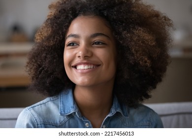 Close up beautiful African teenager girl sitting on sofa at home staring into distance, feel happy, having wide toothy charming smile and natural afro curly hairs posing indoors. Generation Z portrait - Shutterstock ID 2217456013