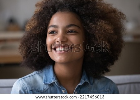 Close up beautiful African teenager girl sitting on sofa at home staring into distance, feel happy, having wide toothy charming smile and natural afro curly hairs posing indoors. Generation Z portrait