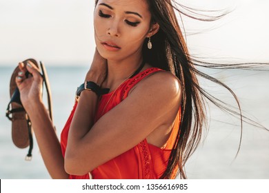 Close Up Of A Beautiful Asian Filipino Girl With Smooth Tan Skin And Her Hair Blowing In The Wind While Holding A Sandal And Wearing An Orange Sleeveless Blouse On The Sandy Beach