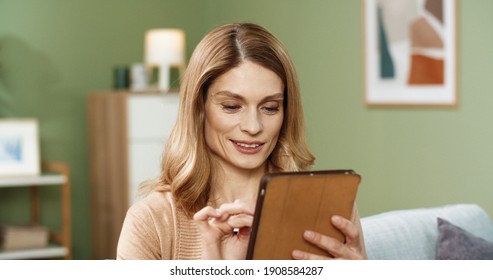 Close Up Of Beautiful Blonde Woman Sitting At Home On Couch And Browsing Catalog Of Goods In Online Store Using Tablet.