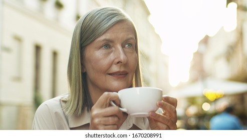 Close Up Of Beautiful Caucasian Senior Woman Wih Gray Hair Drinking Coffee At Cafe Outdoor And Thinking Or Dreaming. Old Lady Sipping Tea While Resting In Center City On Summer Day.