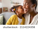 Close up of beautiful daughter kissing mother on cheek at home. African little girl giving kiss to happy mother. Lovely black female child kissing cheerful and proud woman on cheek for mother