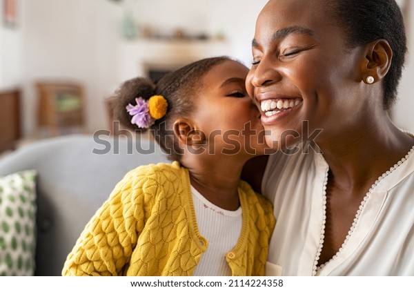 Close up of beautiful daughter kissing mother on\
cheek at home. African little girl giving kiss to happy mother.\
Lovely black female child kissing cheerful and proud woman on cheek\
for mother\'s day.