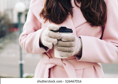 Close up of beautiful female hands in warm wool gloves holding cup of cappuccino coffee. Woman wearing pink coat. Glamorous trendy outfit, hands care, accessories, hot latte, toned instagram colors.