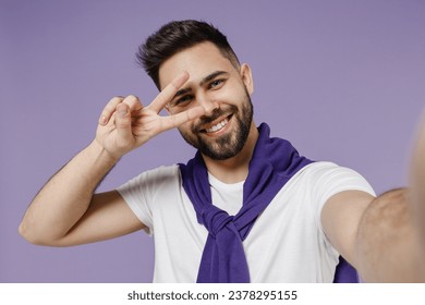 Close up beautiful fun young brunet man 20s wears white t-shirt purple shirt doing selfie shot on mobile cell phone do hide eye behind victory sign isolated on pastel violet background studio portrait