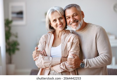 Close Up Of Beautiful Smiling Senior Family Couple Husband And Wife Looking At Camera With Tenderness And Love While Standing In Living Room At Home, Retired Man And Woman Embracing Indoors