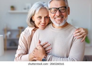 Close Up Of Beautiful Smiling Senior Family Couple Husband And Wife Looking At Camera With Tenderness And Love While Standing In Living Room At Home, Retired Man And Woman Embracing Indoors