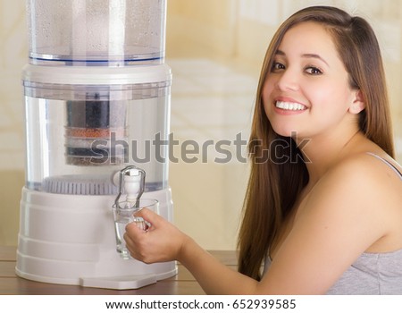 Close up of a beautiful smiling woman filling a glass of water, with a filter system of water purifier on a kitchen background