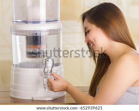 Close up of a beautiful smiling woman filling a glass of water, with a filter system of water purifier on a kitchen background