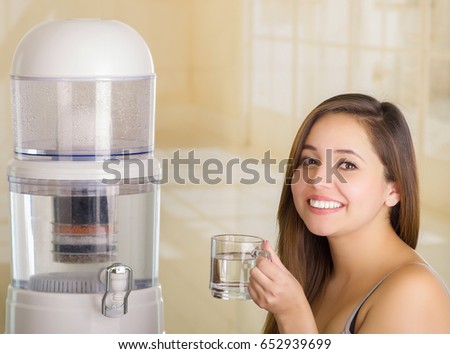 Close up of a beautiful smiling woman holding a glass of water, with a filter system of water purifier on a kitchen background