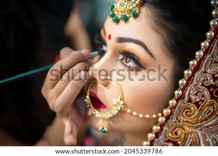 Close up of the beautiful traditional Indian bride getting ready for her wedding day. Cropped hand of makeup artist doing a makeup of bridal face and applying eyeliner.