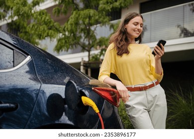 Close up of beautiful woman using smartrphone while charging her electric car on the street.