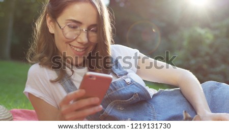 Close up of the beautiful young Caucasian woman using her smartphone and caressing kittycat in the basket while lying and resting in the park.
