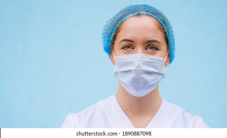 Close Up Of A Beautiful Young Nurse Ready To Work With Surgical Mask On A Light Green Background Looking Straight Into Camera, Smiling