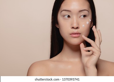 Close up of a beautiful young woman applying moisturizer to her face. Asian female model putting cosmetic cream on her face and looking away.