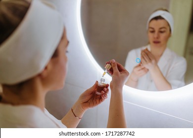 Close Up Of Beautiful Young Woman Applying Cosmetic Oil From A Bottle On Her Face In Bathroom.