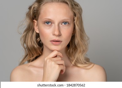 Close up of beautiful young woman face with blue eyes, curly natural blonde hair, has no makeup, touching her chin, standing shirtless with bare shoulders, looking at camera. Studio grey background.
