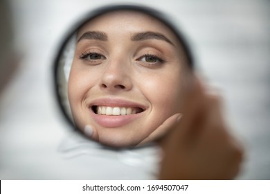 Close Up Of Beautiful Young Woman Look In Mirror Show Healthy White Teeth After Dental Treatment Good Oral Care, Smiling Millennial Female Touch Clean Glowing Face Skin After Beauty Procedures