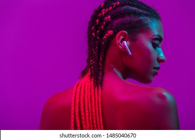 Close up beauty fashion portrait of an attractive young topless african woman wearing dreadlocks standing isolated over purple background, listening to music with earphones