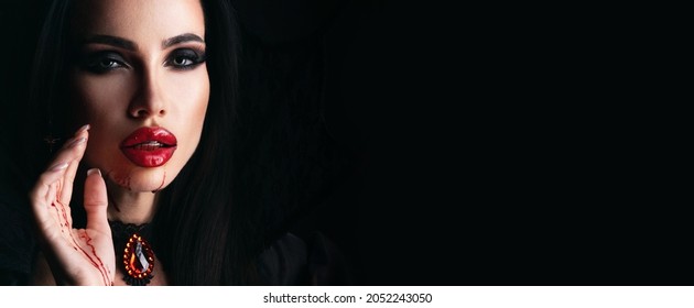 Close up beauty halloween portrait of beautiful young woman as sexy vampire with blood on lips, isolated on black background, banner with copy space for text
