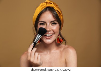 Close up beauty portrait of an attractive lovely young topless woman wearing headband standing isolated over brown background, holding makeup blusher brush - Powered by Shutterstock