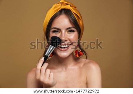 Close up beauty portrait of an attractive lovely young topless woman wearing headband standing isolated over brown background, holding makeup blusher brush