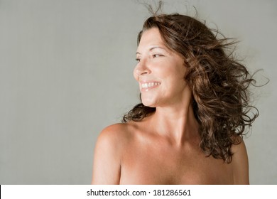 Close Up Beauty Portrait Of An Elegant Mature Woman With Bare Shoulders, Smiling Against A Plain Gray Wall Background While Her Hair Is Flying And Floating In The Breeze. Beauty Lifestyle.