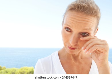 Close Up Beauty Portrait Of A Senior Mature Healthy Woman With Blue Eyes And Flawless Skin Crying And Drying Up Her Tears, Worried And Emotional. Mature And Aging Face With A Sad Expression, Outdoors.