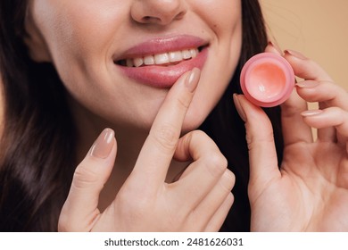 Close up beauty portrait of smiling woman cares of her lips on beige background. Young brunette woman applying make-up cosmetic SPA product. Model using lip balm or gloss for hydration, smoothing