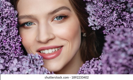 Close up beauty portrait of young beautiful happy smiling woman with healthy flawless radiant skin, natural makeup, posing with blooming lilac flowers. Copy, empty space for text