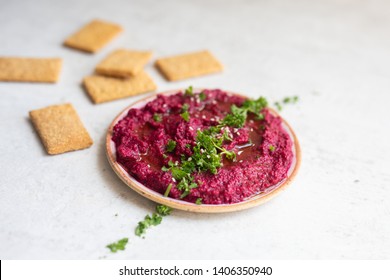 Close up of beet hummus served with hemp seeds, chopped parsley, crackers and a drizzle of olive oil in a pottery bowl on a light natural stone background. Copy space, side view.
