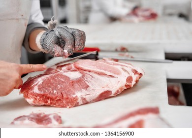 Close up of big fresh chopped pieces of meat and male hands of worker in special gloves. Butcher holding knife and cutting meat pork or beef on table. Manufacturing of raw meat.