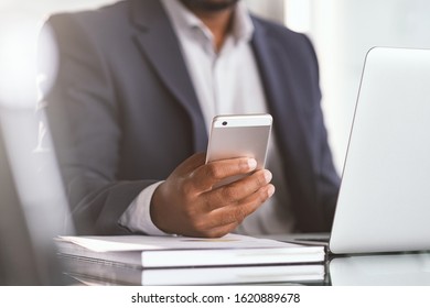 Close Up Of Black Businessman Hand Holding Smartphone While Working On Laptop In Modern Office. Closeup Of African Hand Using Cellphone At Desk. Mature Man Typing On Mobile Phone While Working.