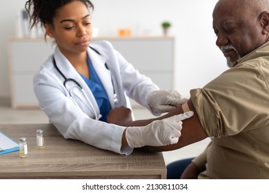 Close Up Of Black Female Doctor In Protective Gloves Applying Sticking Patch To Patient's Arm After Injecting Elderly Man With Seasonal Flu Or Infection Vaccine, Selective Focus On Hand