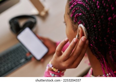 Close up of black teenage girl wearing hearing aid on ear and pressing button while using smartphone at home, copy space - Shutterstock ID 2253204337