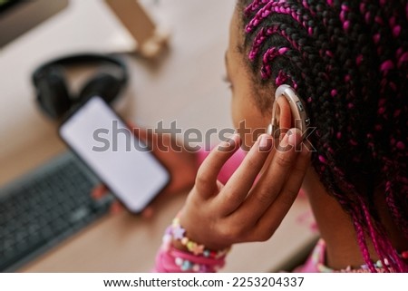 Close up of black teenage girl wearing hearing aid on ear and pressing button while using smartphone at home, copy space