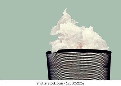 Close up black trash bin with overflowing crumpled paper against a green wall