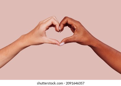 Close up of black and white female hands in heart shape, interracial friendship. Multiracial hands over beige background at studio. Peace and love concept.