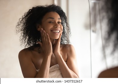 Close up black woman after shower stands wrapped in towel look in mirror touch gentle skin feels happy after cleansing mask, prevention protection, antiaging treatment usage and natural beauty concept