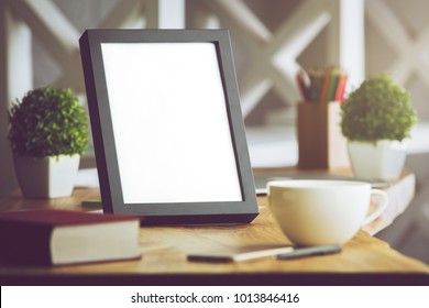 Close Up Of Blank Picture Frame Placed On Office Desk With Coffee Cup, Supplies And Other Items On Blurry Background. Mock Up 