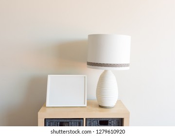 Close up of blank rectangular picture frame with large white lamp on wooden shelf against beige wall