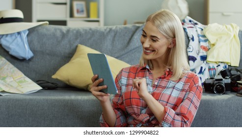 Close Up Of The Blond Charming Young Woman Smiling And Talking Via Web Cam Videochat On The Tablet Computer At Home Among Mess As She Preparing To Go For A Trip.